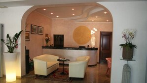 A lobby with a reception desk, two sofas, a small table, and a large potted plant. - Hotel Trifoglio (Jesolo)