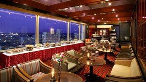 Une chambre d’hôtel située dans un immeuble de grande hauteur, offrant une vue sur la ville, avec un bar et des espaces salon agrémentés de compositions florales.