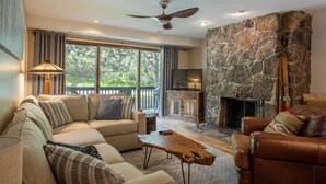 A cozy living room with a stone fireplace, a ceiling fan, and a large window.