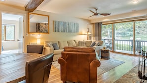 A spacious living area with a large wooden dining table, comfortable seating, a ceiling fan, and a view of the outdoors through large windows.