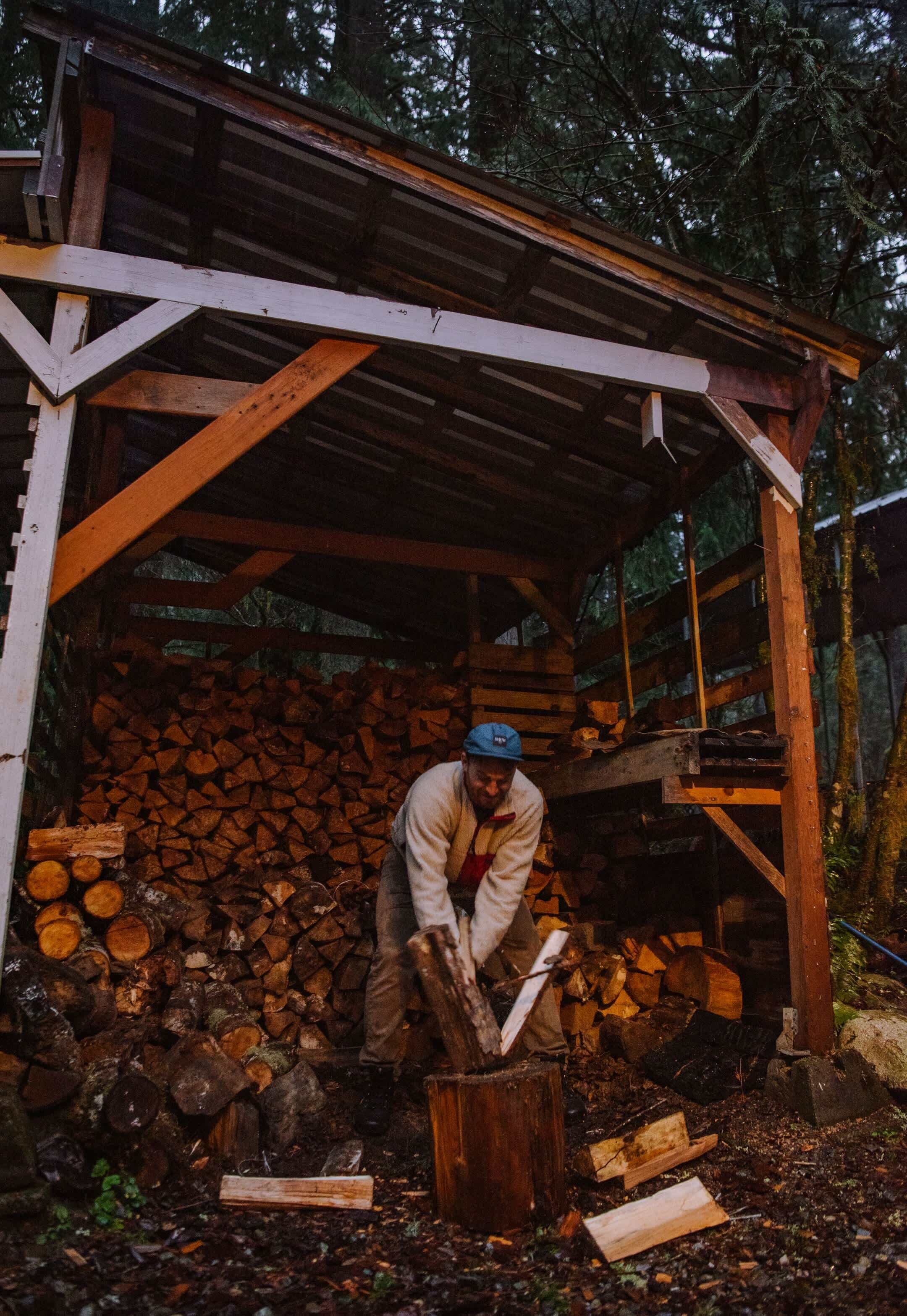 Una persona cortando madera dentro de un rústico cobertizo de madera rodeado de un bosque.