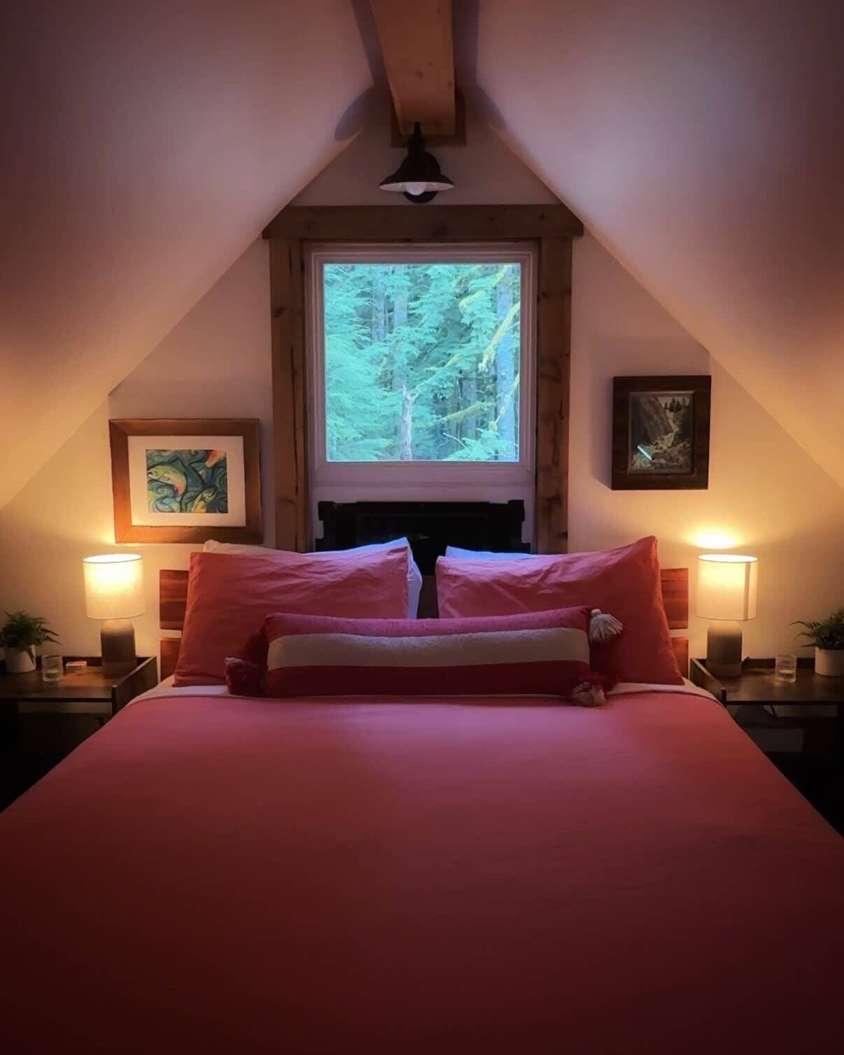 A bedroom with a large bed, two bedside tables with lamps, a window with a forest view, and framed artwork on the walls.