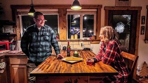 Una mesa de comedor de madera con una talla de ciervo, un hombre y una mujer sentados, una botella y un plato de comida.