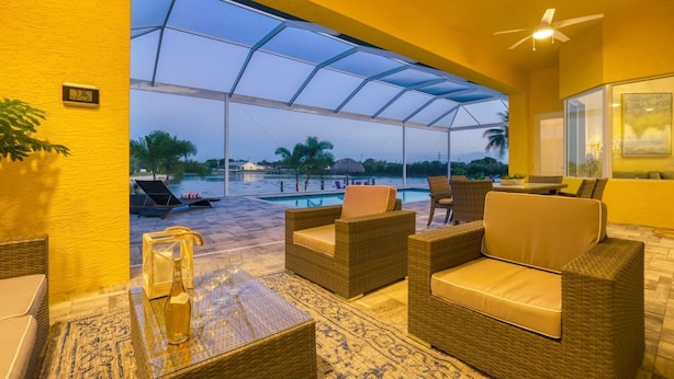 A covered outdoor seating area with wicker furniture and a view of a pool and palm trees. - Full of Sunshine House, Modern, Waterfront w/ Beautiful Heated Pool (Cape Coral)