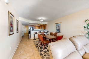 A spacious living room with a beige leather sofa, a dining area with a wooden table and chairs, and a kitchen in the background.