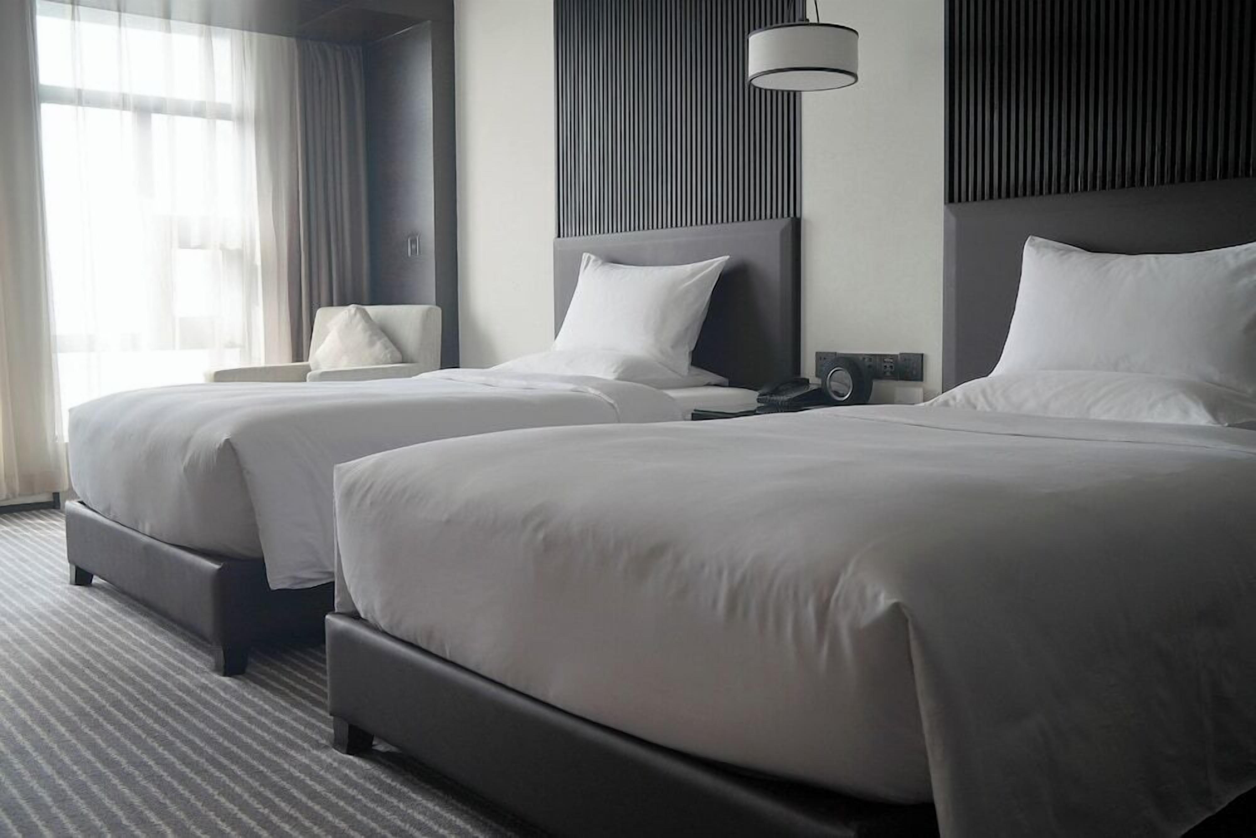 A hotel room with two beds, a bedside table with a lamp, and a window with curtains.