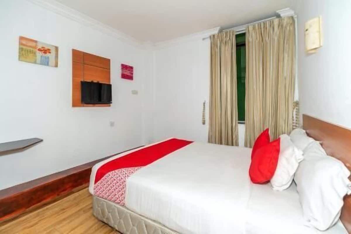 A hotel room with a bed, a wooden nightstand, a red throw blanket, and a window with curtains.