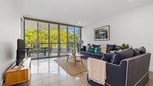 Ein geräumiges Wohnzimmer mit einem großen Schiebeglastür, die zu einem Balkon führt, ein dunkelgraues Sofa mit vielen Kissen, ein Couchtisch aus Holz und ein Fernseher auf einem Holzboden.
