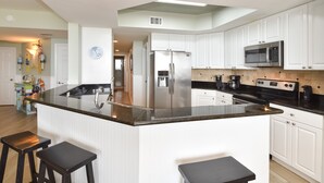 A modern kitchen with white cabinets, a stainless steel refrigerator, a microwave, and a stove. There are two black bar stools at the island.