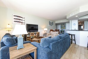 Ein Wohnzimmer mit einem großen, blauen Sofa, einem Holztisch, einem Fernseher und einer Küche im Hintergrund.