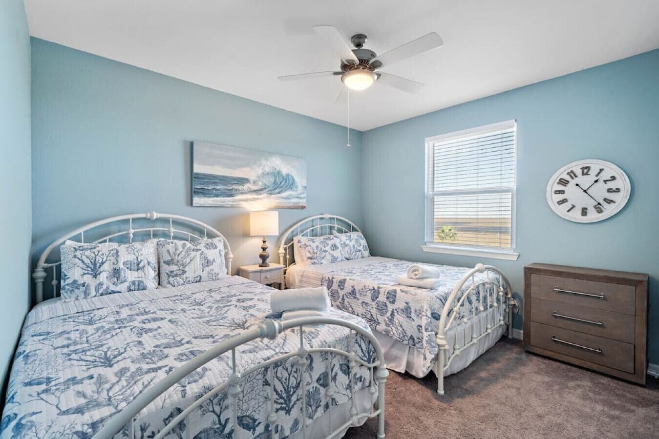 A bedroom with two beds, a ceiling fan, a clock, and a window with blinds.