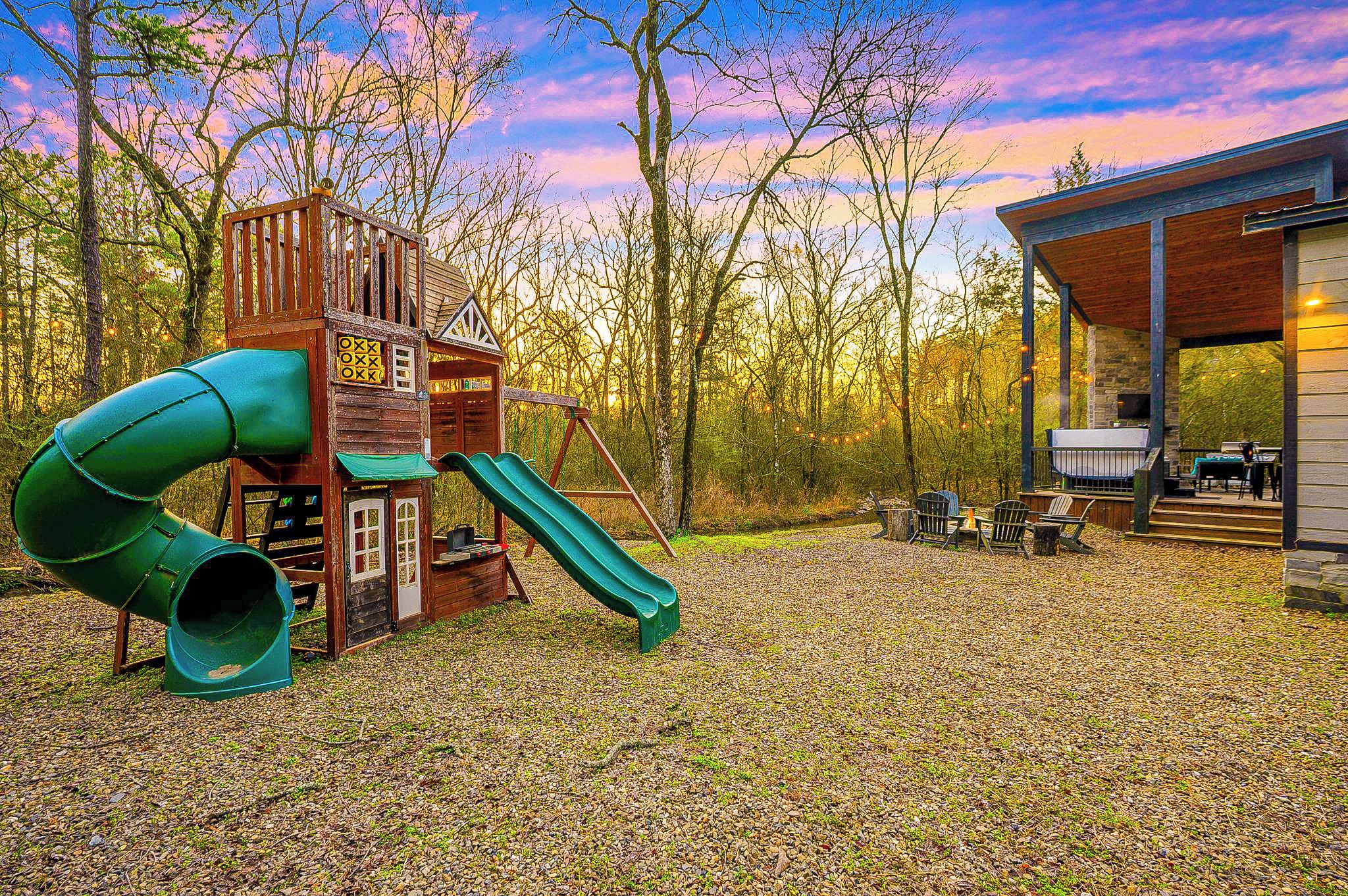 A playground with a slide and climbing structure, surrounded by trees and a building with a covered patio area.