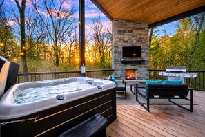 Un bain à remous, un espace salon extérieur, une cheminée en pierre et un barbecue sur une terrasse en bois avec vue sur les arbres.