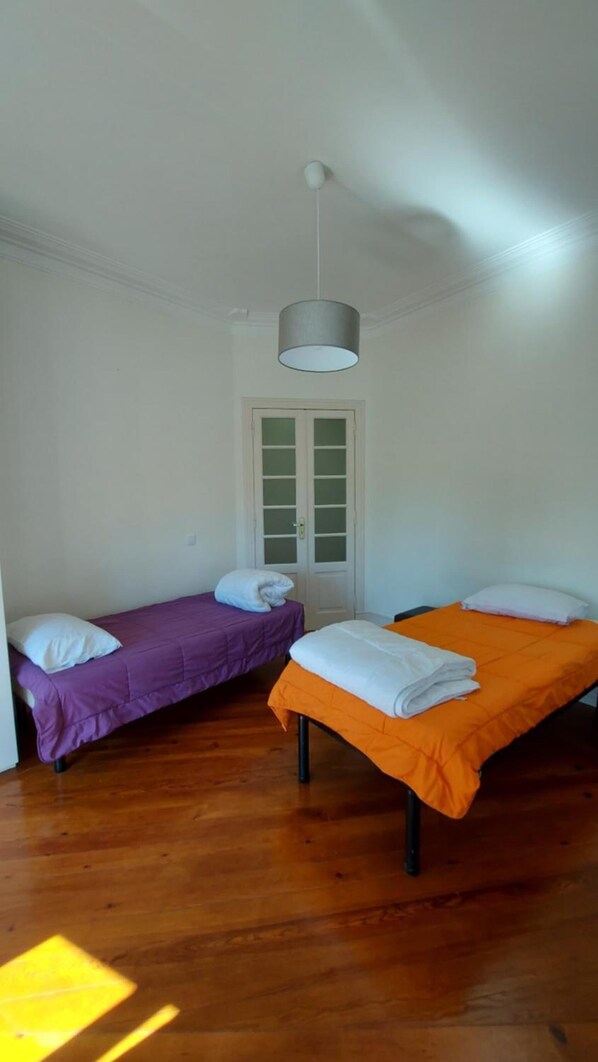 A room with two beds, one with a purple bedspread and the other with an orange bedspread, both with white pillows. There is a white door with frosted glass panels in the background.