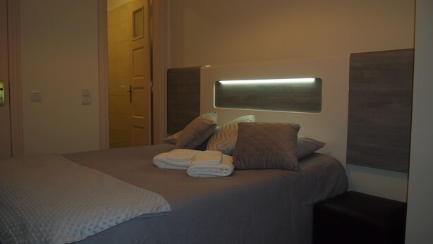 A neatly made bed with pillows and folded towels, a bedside table, and a wall-mounted light fixture. - Green View Home (Lisbon)