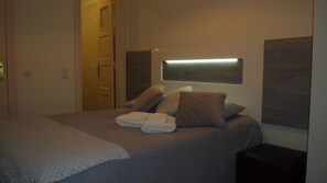 A neatly made bed with pillows and folded towels, a bedside table, and a wall-mounted light fixture.