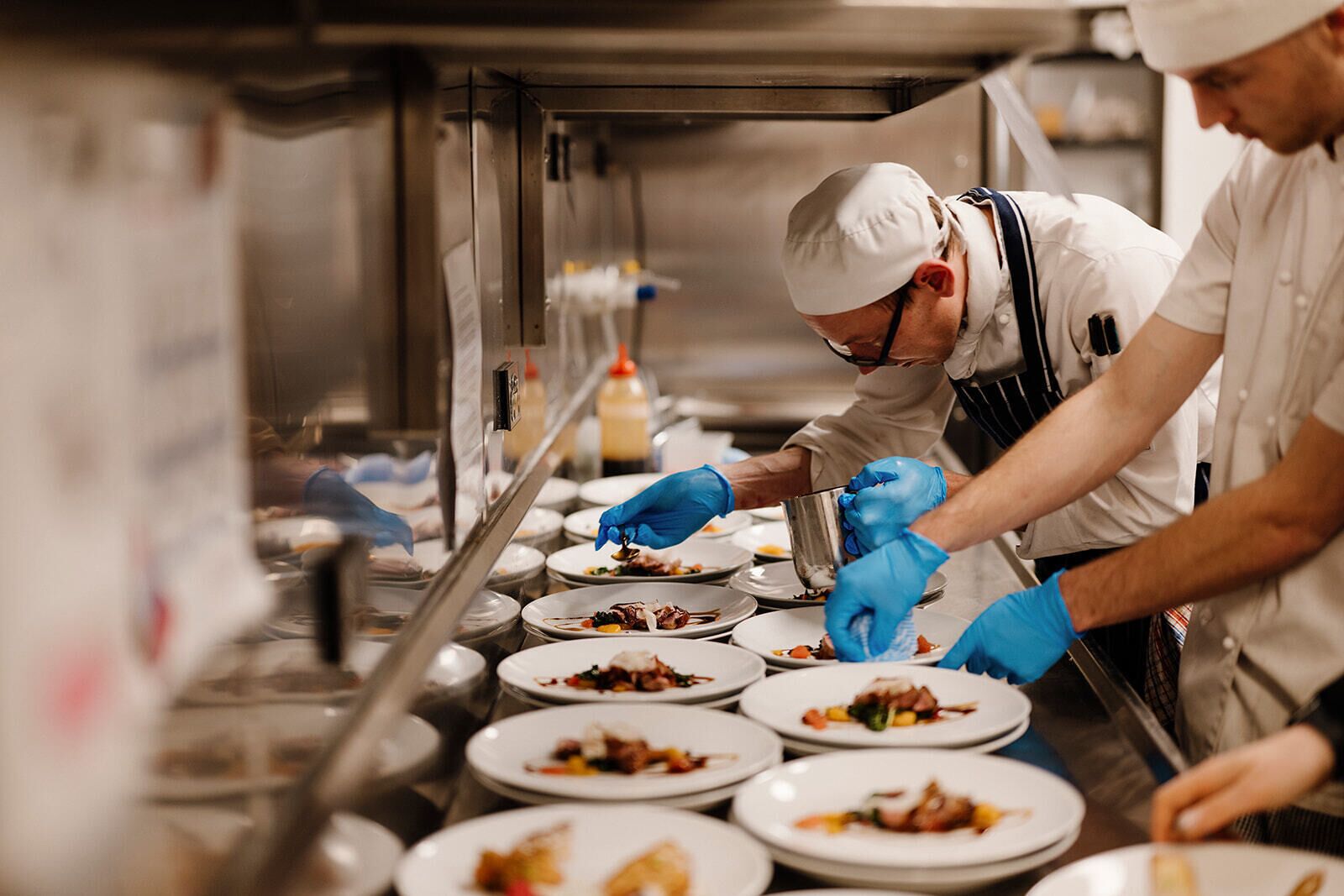 Des chefs préparent des plats dans une cuisine professionnelle dotée de surfaces en acier inoxydable et de divers ustensiles de cuisine.