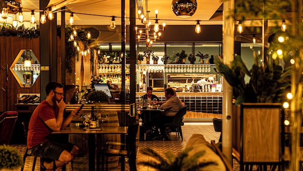 A restaurant with multiple tables and chairs, potted plants, and hanging lights. - The Remington Orange (Orange)