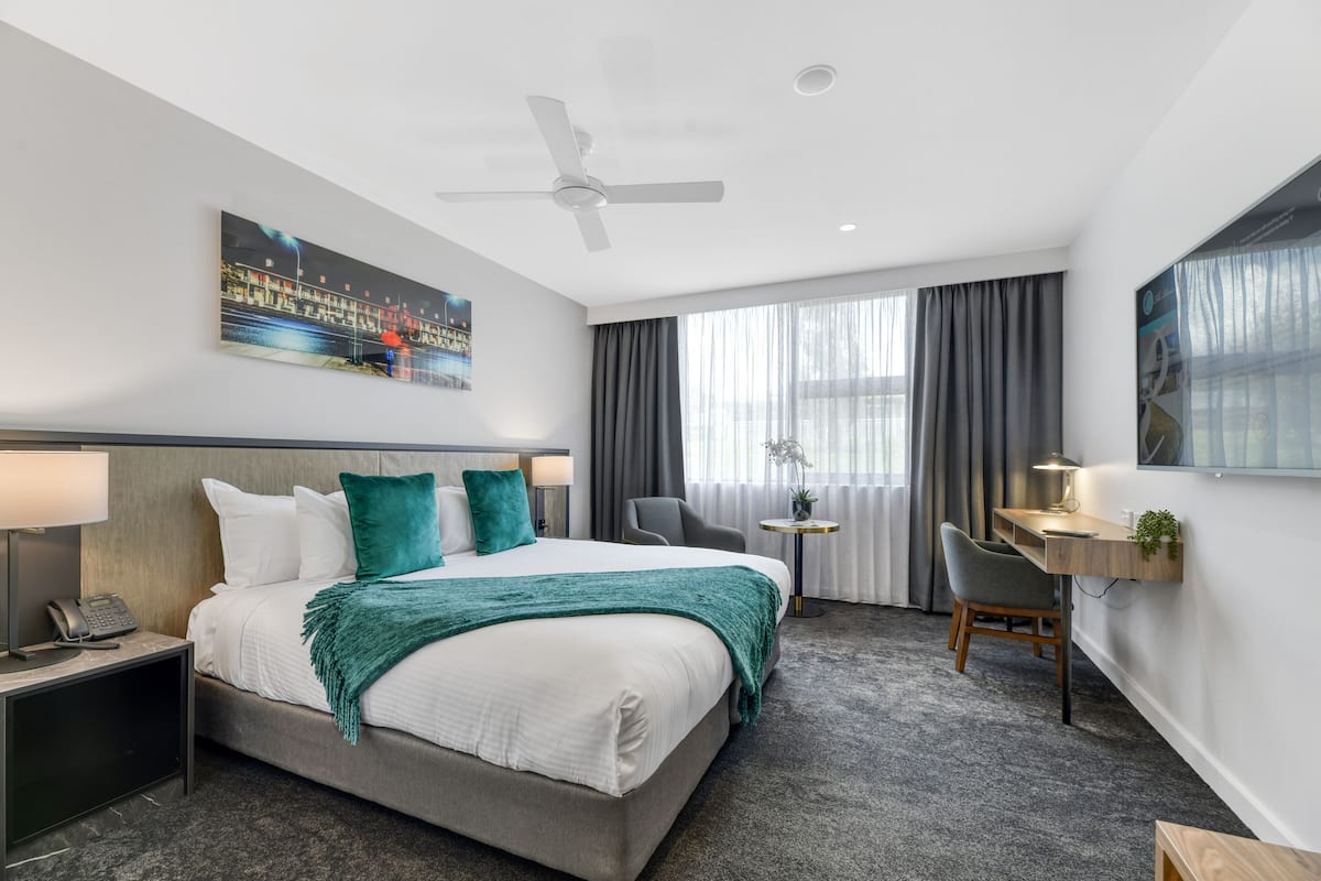 A hotel room with a bed, a desk, a chair, a ceiling fan, and a painting on the wall.
