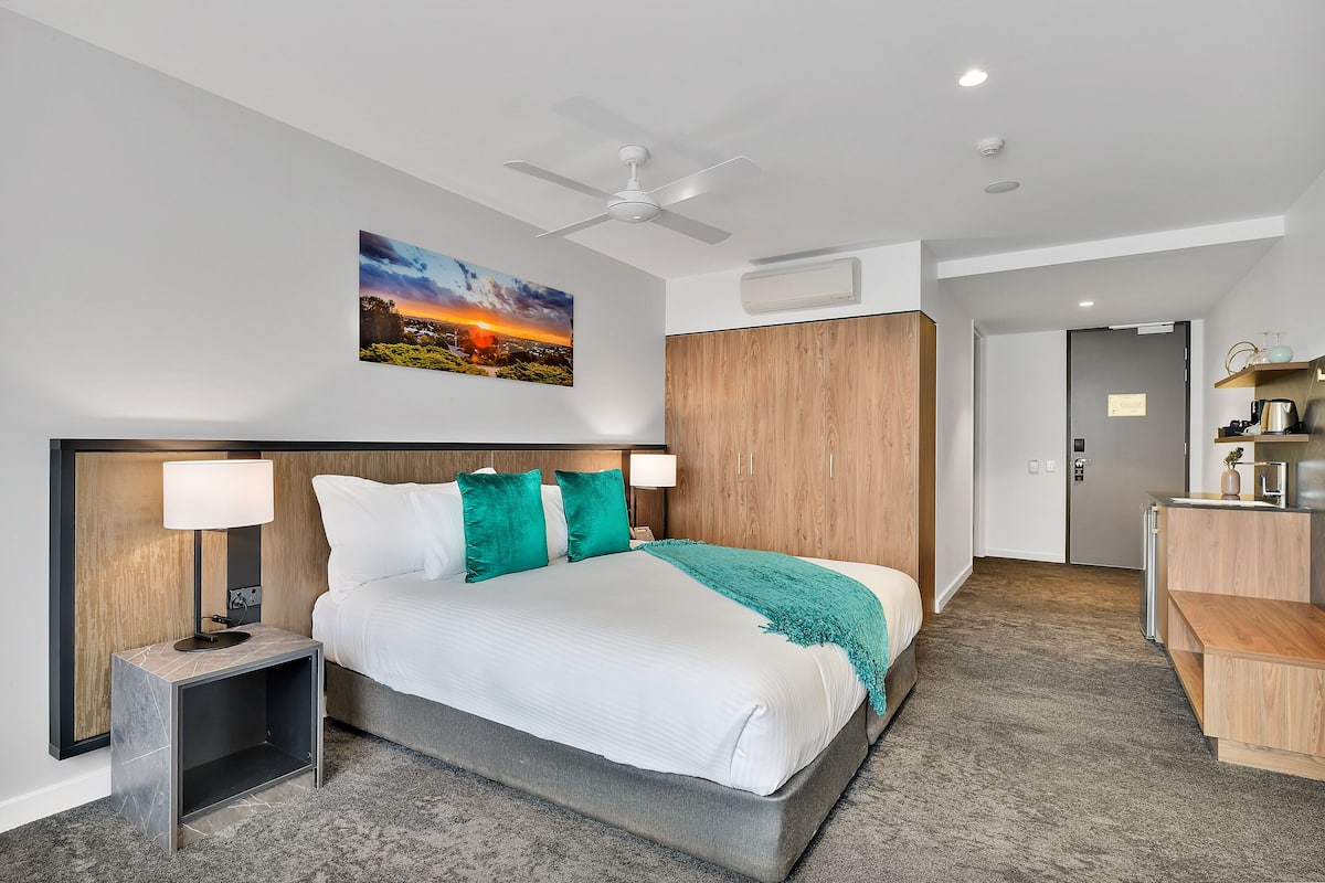 A modern hotel room with a large bed, a nightstand with a lamp, a ceiling fan, a painting of a sunset, a wooden wardrobe, and a kitchenette area.