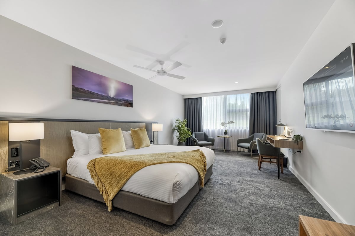 A hotel room with a large bed, a desk, a chair, a small table, a plant, and a painting on the wall.