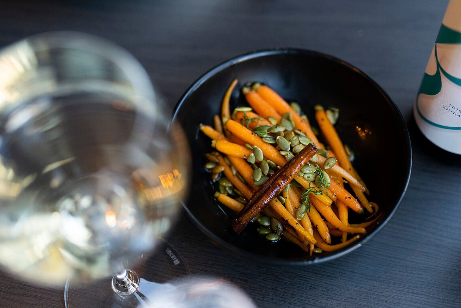 Un tazón de zanahorias asadas con semillas de calabaza y una ramita de canela, acompañado de una copa de vino blanco sobre una mesa de madera.