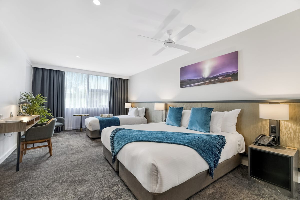 A hotel room with two beds, a desk, a chair, a small table, a plant, and a painting on the wall.
