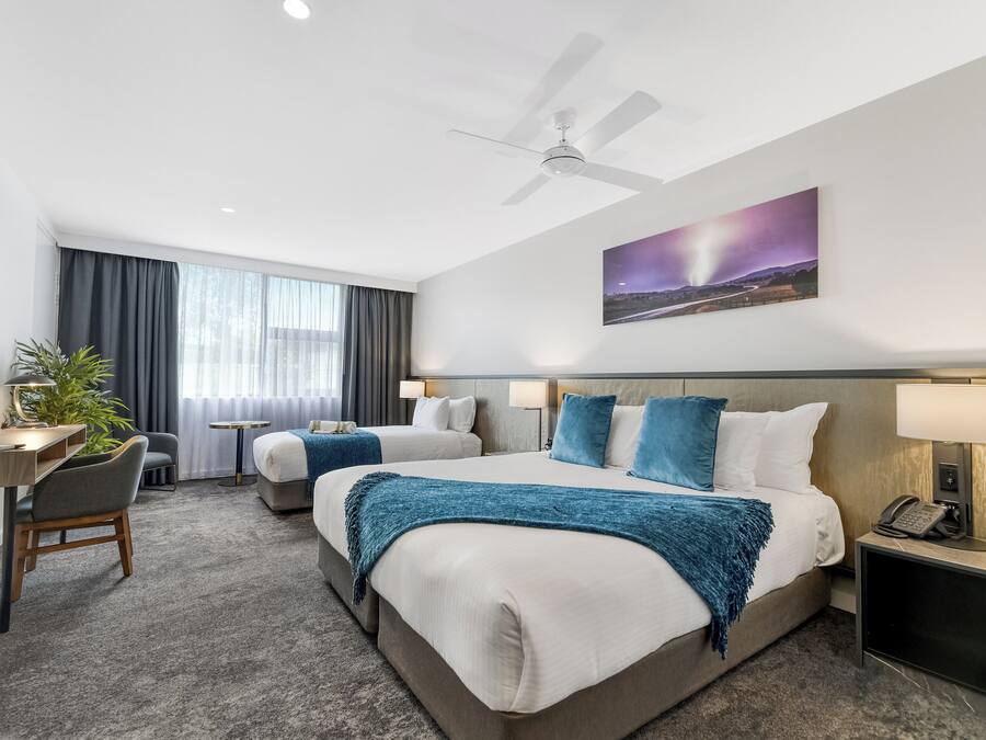 A hotel room with two beds, a desk, a chair, a small table, a plant, and a painting on the wall.