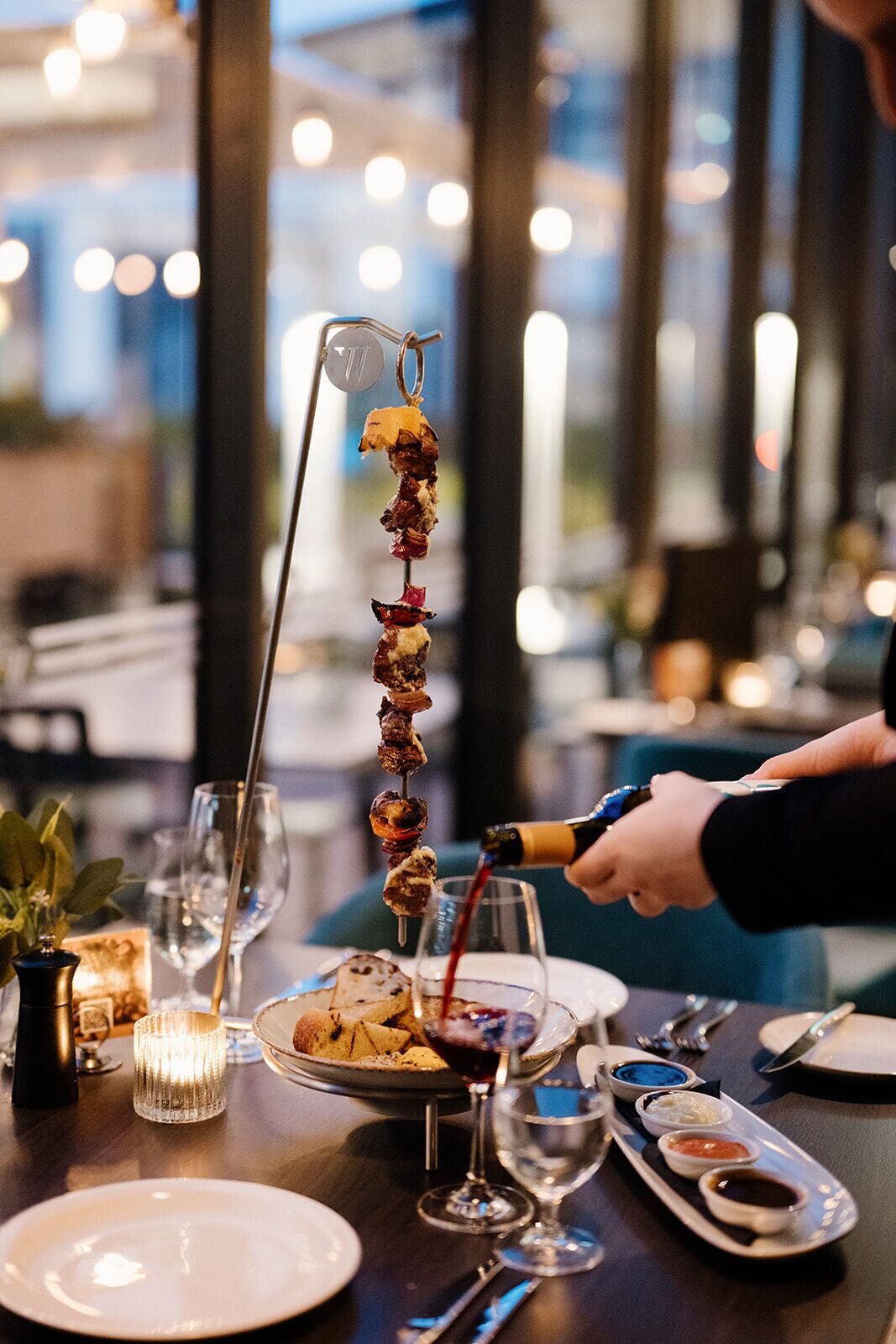 Une table à manger avec un plat de viande grillée, une bouteille de vin rouge dont on verse le contenu, ainsi que diverses assiettes et verres.