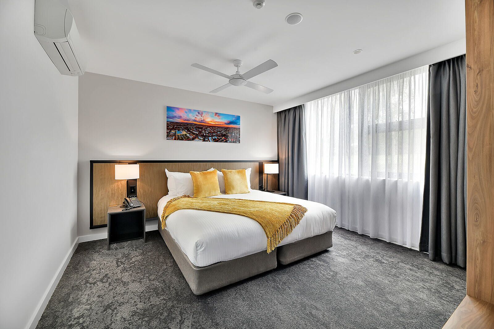 Une chambre d’hôtel avec un grand lit, un ventilateur de plafond, une table de chevet avec une lampe, un tableau au mur et une vue sur l’extérieur à travers des rideaux légers.