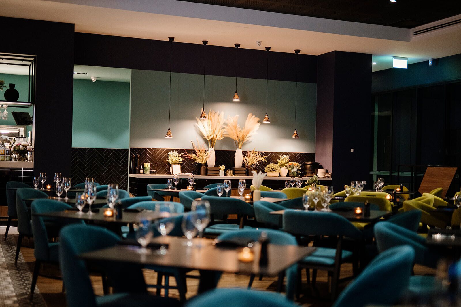 Un interior de restaurante moderno con sillas color turquesa, mesas dispuestas con copas de vino y plantas decorativas.