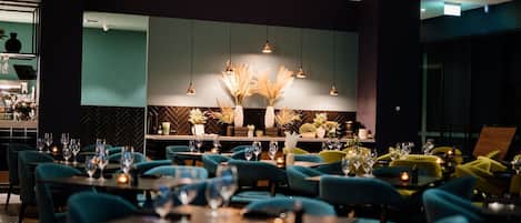 Un interior de restaurante moderno con sillas color turquesa, mesas dispuestas con copas de vino y plantas decorativas.