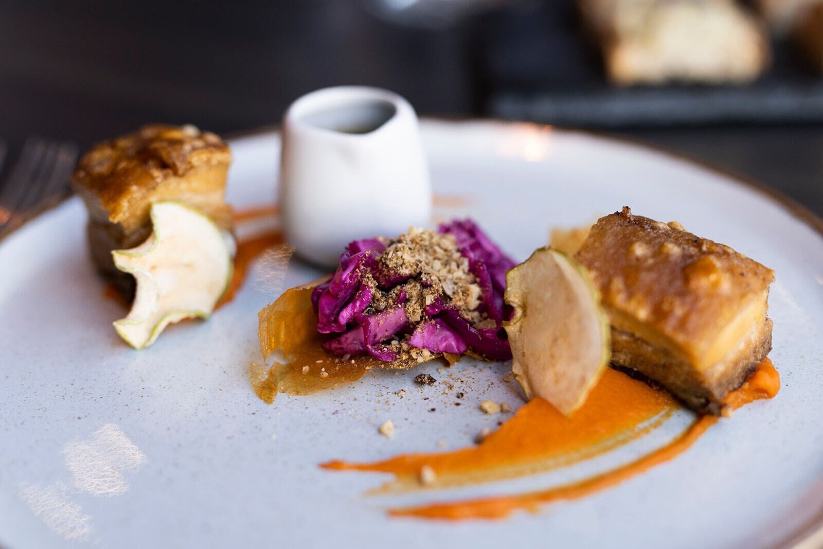 Un plato con un trozo de carne, una guarnición de col morada y una taza blanca pequeña.