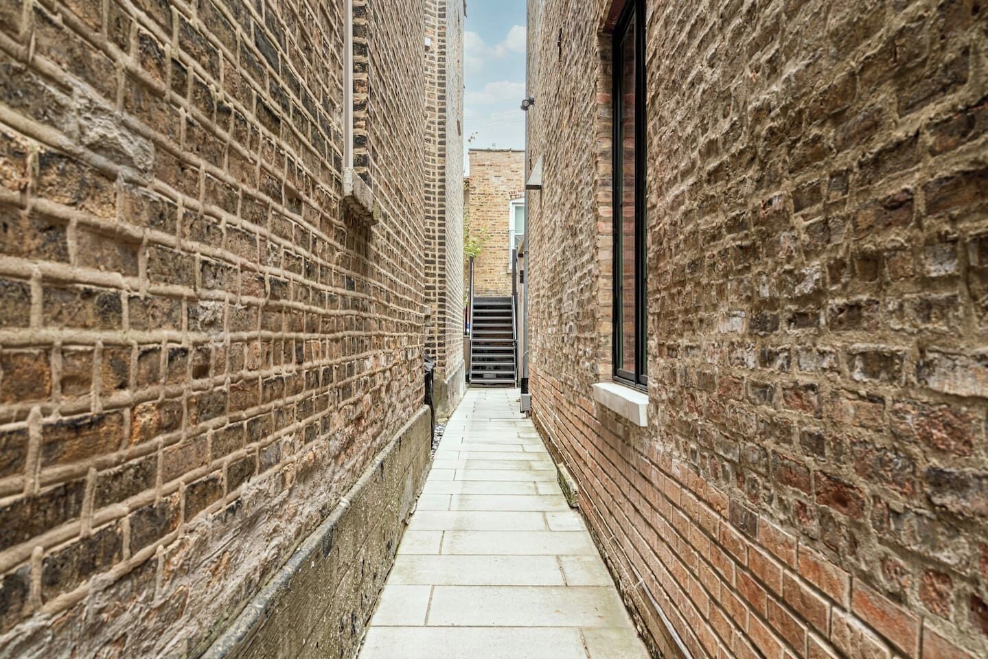 A narrow alleyway with brick walls and a tiled floor, leading to a gate.