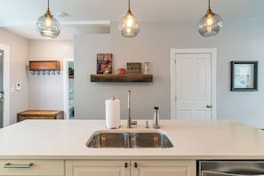 A kitchen with a large island, stainless steel sink, hanging pendant lights, and a wooden shelf with decorative items.