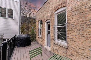 Una terraza de madera con una parrilla, un banco y un edificio de ladrillo con ventanas.