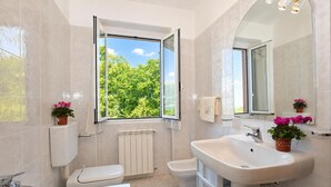 Ein Badezimmer mit Toilette, Bidet, Badewanne und Waschbecken, mit einem Fenster mit Blick auf Grünflächen.