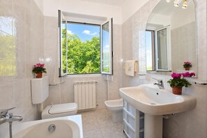 A bathroom with a toilet, bidet, bathtub, and sink, with a window offering a view of greenery. - Viola (Colico)
