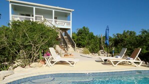 Un área de piscina con sillones de descanso, una escalera que lleva a una casa y follaje circundante.