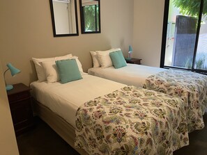 Une chambre d’hôtel avec deux lits, chacun recouvert d’une courtepointe blanche et agrémenté d’un oreiller turquoise. On y trouve deux tables de chevet avec des lampes, une fenêtre donnant sur un paysage verdoyant et un miroir accroché au mur.