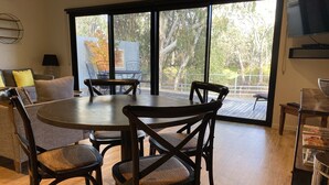 A dining area with a round table and chairs, large windows with a view of trees, a sofa with cushions, and a TV mounted on the wall. - Adelphi 3 - River views located in the centre of Echuca (Echuca)