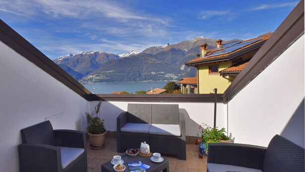 A balcony with wicker furniture, a table with breakfast items, and a scenic mountain view. - Belvedere A (Colico)