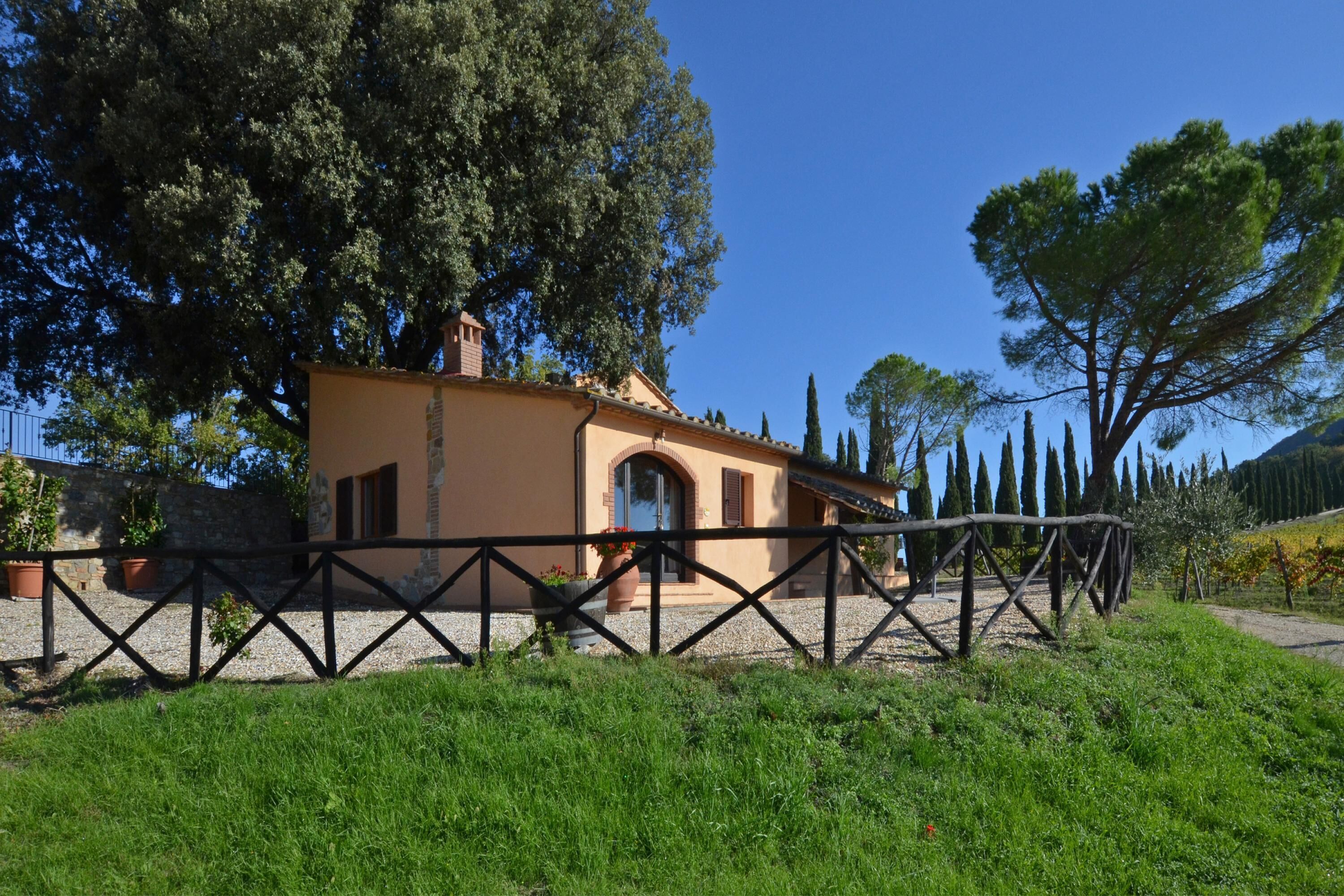 A rustic house with a stone wall, a wooden fence, and a large tree in the background.