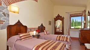 A bedroom with two wooden beds, a stone wall, a mirror, and a window with a view of trees. - Paradiso Villas (Casole d'Elsa)