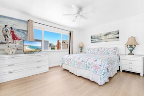 A bedroom with a bed, a TV, a ceiling fan, a window with curtains, and a painting on the wall.