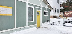 Un edificio con un letrero de alquiler vacacional, una puerta amarilla y bancos cubiertos de nieve.