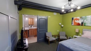 Une chambre à coucher avec un lit, deux fauteuils à motifs, un bureau et, au fond, une petite cuisine.