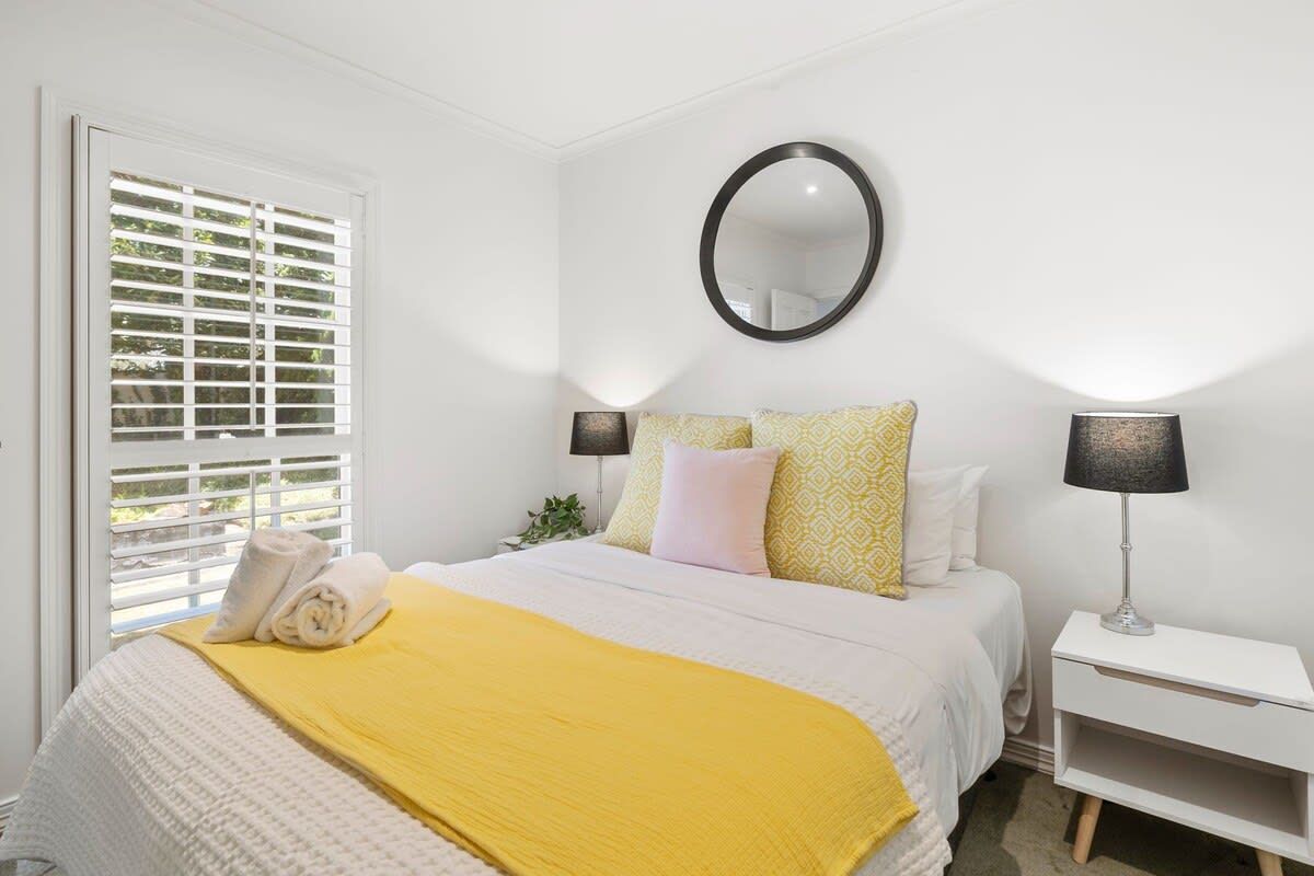 Une chambre à coucher avec un lit, une table de chevet, une lampe, un miroir et une fenêtre avec des stores.