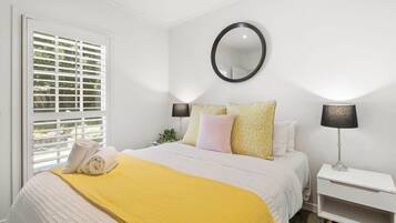 Une chambre à coucher avec un lit, une table de chevet, une lampe, un miroir et une fenêtre avec des stores.