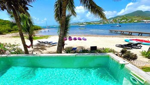 A pool with clear water, surrounded by lounge chairs and a sandy beach with boats in the background.